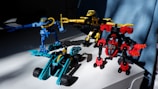 Three colorful toy robots are positioned on a white surface. The robots are constructed with interlocking plastic pieces in blue, yellow, and red colors. Sunlight casts shadows, adding depth to the scene, and a blue curtain is visible in the background.