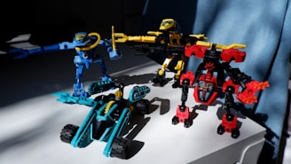 Three colorful toy robots are positioned on a white surface. The robots are constructed with interlocking plastic pieces in blue, yellow, and red colors. Sunlight casts shadows, adding depth to the scene, and a blue curtain is visible in the background.