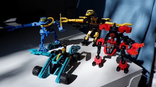 Three colorful toy robots are positioned on a white surface. The robots are constructed with interlocking plastic pieces in blue, yellow, and red colors. Sunlight casts shadows, adding depth to the scene, and a blue curtain is visible in the background.