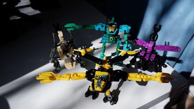 Four colorful toy robot figures arranged on a white surface, each in different postures with bright lighting casting shadows on some parts. The toys are primarily composed of pieces in bright yellow, turquoise, purple, and beige.