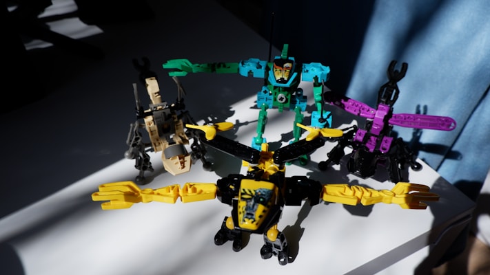 Four colorful toy robot figures arranged on a white surface, each in different postures with bright lighting casting shadows on some parts. The toys are primarily composed of pieces in bright yellow, turquoise, purple, and beige.