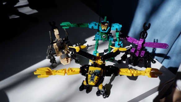 Four colorful toy robot figures arranged on a white surface, each in different postures with bright lighting casting shadows on some parts. The toys are primarily composed of pieces in bright yellow, turquoise, purple, and beige.