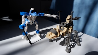 Two robotic figures constructed from interlocking blocks are positioned on a flat surface. One figure is primarily blue and white, resembling a humanoid with a stick-like appendage, while the other is tan and gray with a mechanical, quadruped appearance. The sunlight casts strong shadows, creating a dynamic contrast.