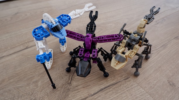 Three robot-like figures made from interlocking building blocks are arranged on a wooden surface. Each robot is distinctly colored, with one primarily in blue and white, another in black and purple, and the third in tan and black. The structures resemble creatures with multiple legs.
