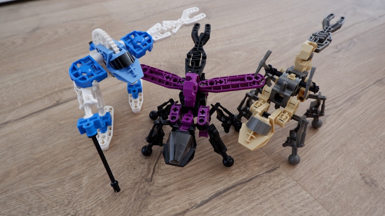 Three robot-like figures made from interlocking building blocks are arranged on a wooden surface. Each robot is distinctly colored, with one primarily in blue and white, another in black and purple, and the third in tan and black. The structures resemble creatures with multiple legs.