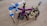 Three robot-like figures made from interlocking building blocks are arranged on a wooden surface. Each robot is distinctly colored, with one primarily in blue and white, another in black and purple, and the third in tan and black. The structures resemble creatures with multiple legs.
