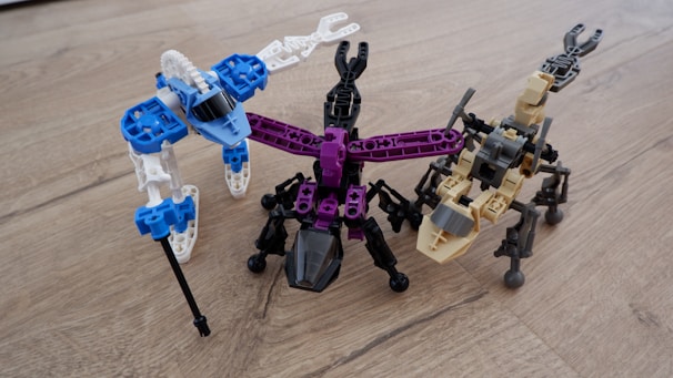 Three robot-like figures made from interlocking building blocks are arranged on a wooden surface. Each robot is distinctly colored, with one primarily in blue and white, another in black and purple, and the third in tan and black. The structures resemble creatures with multiple legs.