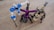 Three robot-like figures made from interlocking building blocks are arranged on a wooden surface. Each robot is distinctly colored, with one primarily in blue and white, another in black and purple, and the third in tan and black. The structures resemble creatures with multiple legs.