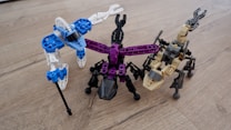 Three robot-like figures made from interlocking building blocks are arranged on a wooden surface. Each robot is distinctly colored, with one primarily in blue and white, another in black and purple, and the third in tan and black. The structures resemble creatures with multiple legs.