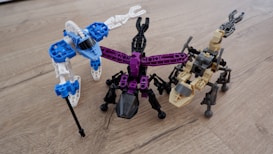 Three robot-like figures made from interlocking building blocks are arranged on a wooden surface. Each robot is distinctly colored, with one primarily in blue and white, another in black and purple, and the third in tan and black. The structures resemble creatures with multiple legs.
