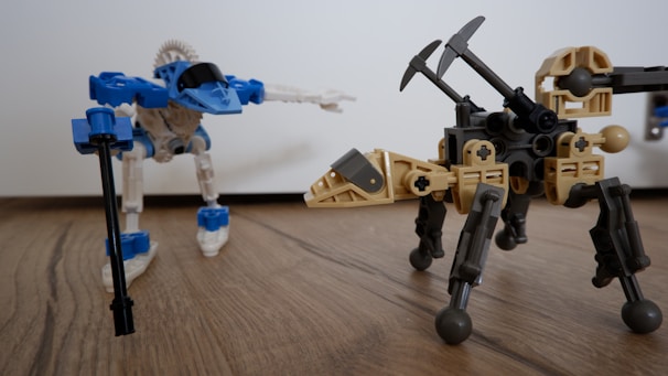 A scene featuring two robotic figures made from interlocking pieces on a wooden surface. One robot is primarily blue and white with a futuristic design, holding a black rod in one hand. The other robot is tan and black with insect-like legs and two pickaxe arms, resembling a mechanical creature.