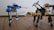 A scene featuring two robotic figures made from interlocking pieces on a wooden surface. One robot is primarily blue and white with a futuristic design, holding a black rod in one hand. The other robot is tan and black with insect-like legs and two pickaxe arms, resembling a mechanical creature.