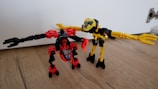 Two toy robots feature prominently, crafted from colorful plastic pieces. One is predominantly red and black, with a shield-like emblem on its chest and a small fire piece in its hand. The other is primarily yellow and black, with extended arms resembling mechanical forks. They stand on a wooden floor with a white wall and part of a door hinge visible in the background.