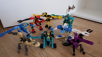 Several colorful Bionicle-like toys are posed on a wooden floor, each made of various interlocking parts. The figures include a range of colors like red, blue, green, and purple, with distinct robotic and creature-like designs. The toys are arranged in a semi-circle, each exhibiting different stances and features, showcasing a playful and imaginative setup.