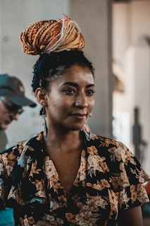 A person is standing indoors, wearing a floral-patterned outfit. Her hair is styled in an intricate updo with a mix of brown, orange, and white tones. She is wearing long earrings and has a calm expression on her face.