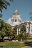 A large neoclassical government building with a prominent dome surrounded by lush greenery, benches, and a flagpole. The architecture features pillars and intricate design elements typical of a capitol building. The bright sky and trees enhance the setting.