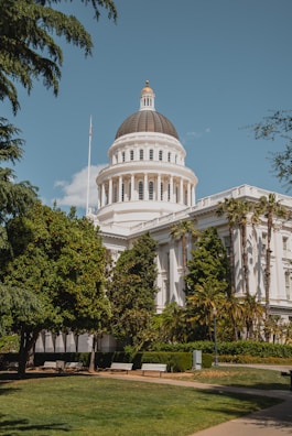 A large neoclassical government building with a prominent dome surrounded by lush greenery, benches, and a flagpole. The architecture features pillars and intricate design elements typical of a capitol building. The bright sky and trees enhance the setting.