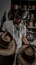 A person wearing a top hat and a t-shirt is looking down while holding two wide-brimmed hats. Behind them is a shelf filled with various hats and objects.