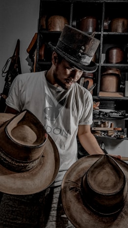 A person wearing a top hat and a t-shirt is looking down while holding two wide-brimmed hats. Behind them is a shelf filled with various hats and objects.