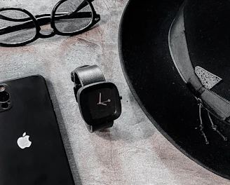 A sleek arrangement of personal items consisting of a classic black fedora hat, an Apple smartphone, a pair of round eyeglasses, and a modern black smartwatch. The items are positioned on a textured stone surface, creating a minimalist and stylish composition.
