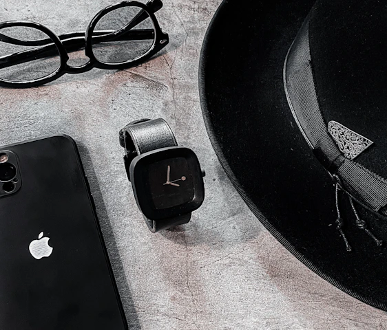 A sleek arrangement of personal items consisting of a classic black fedora hat, an Apple smartphone, a pair of round eyeglasses, and a modern black smartwatch. The items are positioned on a textured stone surface, creating a minimalist and stylish composition.