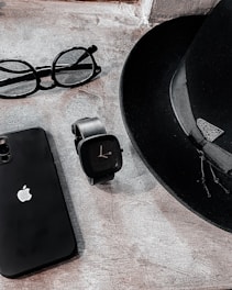 A sleek arrangement of personal items consisting of a classic black fedora hat, an Apple smartphone, a pair of round eyeglasses, and a modern black smartwatch. The items are positioned on a textured stone surface, creating a minimalist and stylish composition.