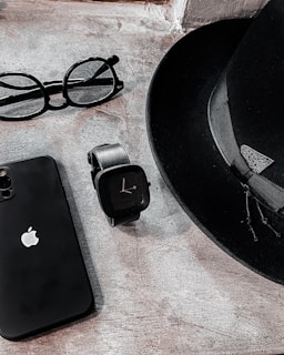 A sleek arrangement of personal items consisting of a classic black fedora hat, an Apple smartphone, a pair of round eyeglasses, and a modern black smartwatch. The items are positioned on a textured stone surface, creating a minimalist and stylish composition.