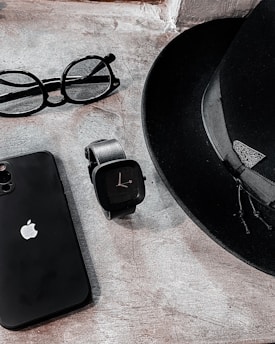 A sleek arrangement of personal items consisting of a classic black fedora hat, an Apple smartphone, a pair of round eyeglasses, and a modern black smartwatch. The items are positioned on a textured stone surface, creating a minimalist and stylish composition.