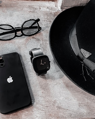 A sleek arrangement of personal items consisting of a classic black fedora hat, an Apple smartphone, a pair of round eyeglasses, and a modern black smartwatch. The items are positioned on a textured stone surface, creating a minimalist and stylish composition.