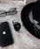 A sleek arrangement of personal items consisting of a classic black fedora hat, an Apple smartphone, a pair of round eyeglasses, and a modern black smartwatch. The items are positioned on a textured stone surface, creating a minimalist and stylish composition.