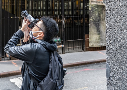 A person wearing a face mask and sunglasses is taking a photograph with a large camera. They are standing on a street, facing away from the camera. The person has short, dark hair and is wearing a black jacket with a backpack. In the background, there is a metal fence and a poster partially visible. The side of a building is also seen in the foreground.