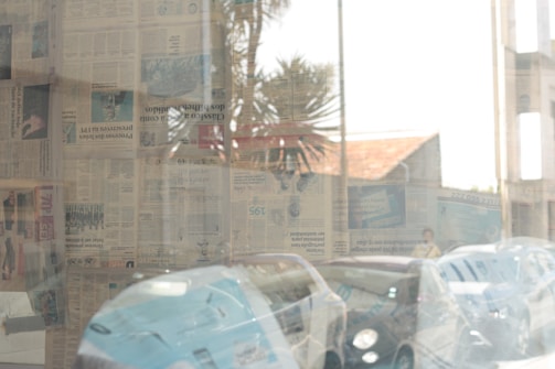 A series of newspapers covers a glass window, creating a collage effect with various headlines and images visible. Reflections in the glass show some cars parked outside and elements of the surrounding environment, including a palm tree and part of a building with a tiled roof.