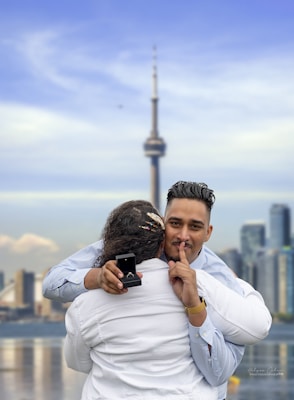 A man with a gentle smile holds a ring box while embracing a person dressed in white, suggesting an engagement or proposal. The man has a finger to his lips, indicating a gesture of secrecy or surprise. The background features a prominent tower and skyscrapers, indicating an urban setting.