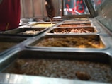 A variety of steaming dishes are presented in stainless steel trays, with someone holding a ladle over one of them. The food appears rich in color and texture, indicating a diverse selection.