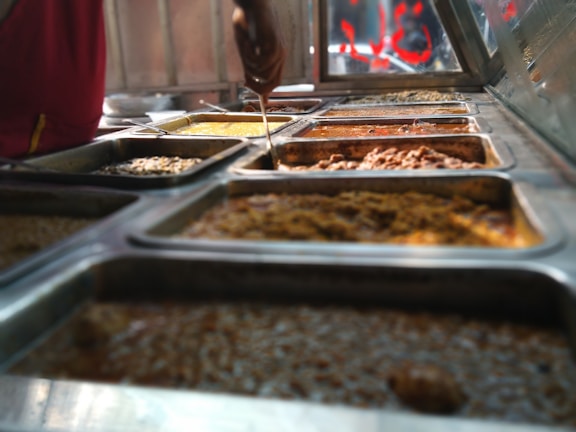 A variety of steaming dishes are presented in stainless steel trays, with someone holding a ladle over one of them. The food appears rich in color and texture, indicating a diverse selection.