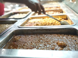 Several trays of food are displayed in a cafeteria-style setting, with different types of dishes in each tray. A hand holding a ladle is seen serving food from one of the trays, filled with a yellowish sauce. The closest tray in the foreground contains a dark, chunky stew-like dish.