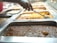 Several trays of food are displayed in a cafeteria-style setting, with different types of dishes in each tray. A hand holding a ladle is seen serving food from one of the trays, filled with a yellowish sauce. The closest tray in the foreground contains a dark, chunky stew-like dish.