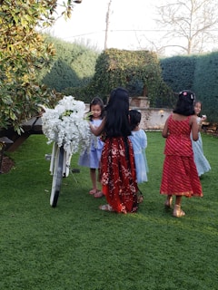 A group of children in colorful dresses gather around a white bicycle adorned with white flowers, set in a lush green garden with tall hedges and trees in the background. They appear to be engaged in a playful moment together.