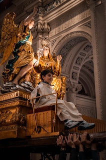 A ceremonial religious procession is depicted, featuring a golden statue of a religious figure adorned with elaborate garments and a crown, positioned beside a golden angel figurine. A young boy dressed in white robes is seated on a decorated wooden platform carried by several hands from individuals positioned below the frame. The intricate architectural background enhances the solemnity and grandeur of the scene.