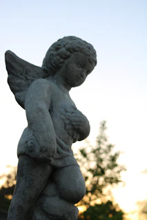 A stone sculpture of an angel with wings is set against a backdrop of a soft, fading sunset. The sculpture features intricate details on the angel’s hair and wings, with a serene expression on its face. The background shows blurred foliage silhouetted by the warm glow of a sunset.