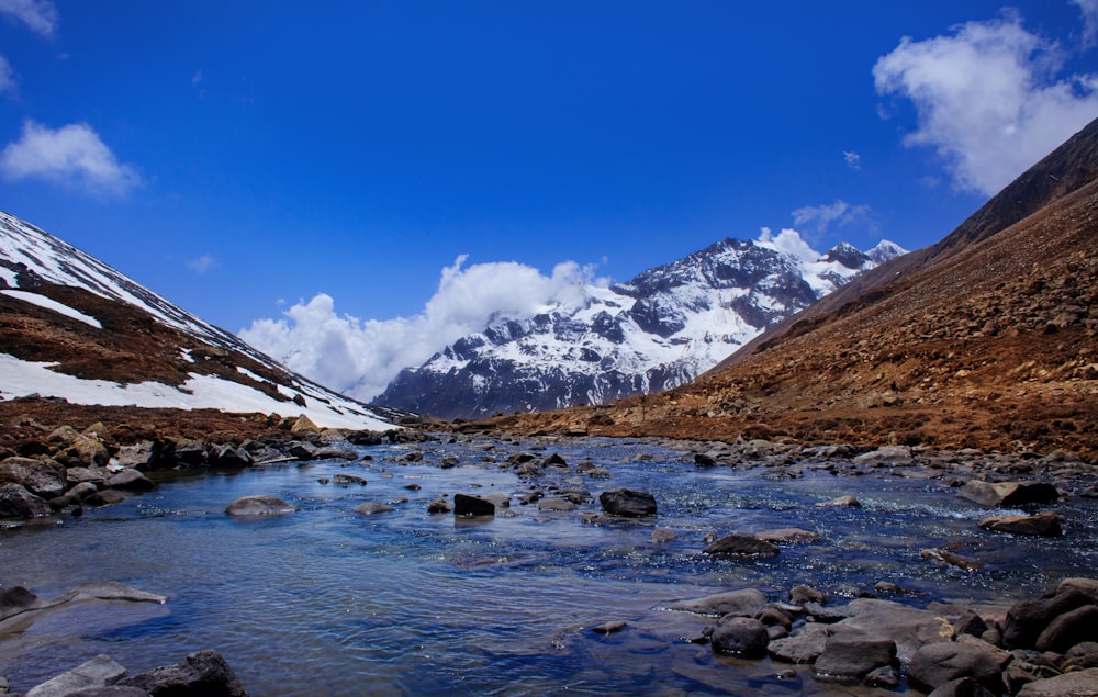 a stream running through a rocky valley with snow covered mountains in the background