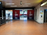 An indoor corridor leading to an entrance with G20 Indonesia 2022 signage prominently displayed. The area is well-lit with overhead fluorescent lights and has a tiled floor. A railing runs along the left side, and there is a sign for arrivals on the right. In the background, there is a glimpse of an open area with seating and people moving around.