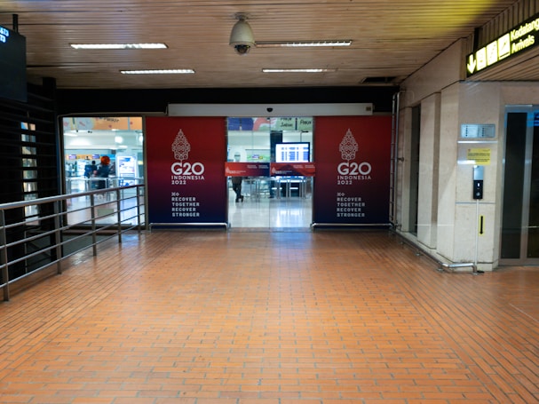 An indoor corridor leading to an entrance with G20 Indonesia 2022 signage prominently displayed. The area is well-lit with overhead fluorescent lights and has a tiled floor. A railing runs along the left side, and there is a sign for arrivals on the right. In the background, there is a glimpse of an open area with seating and people moving around.
