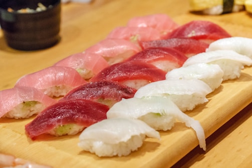 An assortment of sushi neatly arranged on a wooden serving board, featuring pieces with different types of fish such as tuna and white fish atop small mounds of rice. The setting suggests a clean, well-prepared presentation typical of Japanese cuisine.