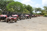 A row of all-terrain vehicles (ATVs) parked on a gravel surface under a clear sky, surrounded by lush green trees and palm trees. The ATVs have a red color and show signs of being used off-road, with some dirt accumulation on their tires and bodies.
