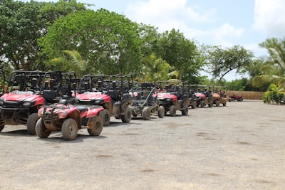 A row of all-terrain vehicles (ATVs) parked on a gravel surface under a clear sky, surrounded by lush green trees and palm trees. The ATVs have a red color and show signs of being used off-road, with some dirt accumulation on their tires and bodies.