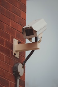 A surveillance camera mounted on a red brick wall. The camera is attached to a beige metal arm and is facing outward. Below the camera is an electrical junction box connected by a black cable.