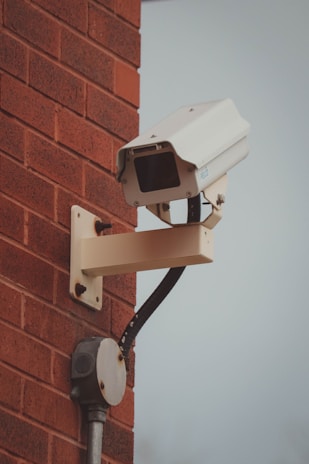 A surveillance camera mounted on a red brick wall. The camera is attached to a beige metal arm and is facing outward. Below the camera is an electrical junction box connected by a black cable.
