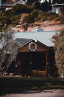 A quaint wooden building nestled among dense greenery features a life preserver with the words 'Canadian Bay Club' displayed prominently at the front. The structure has a small porch with rustic railings, surrounded by trees and shrubs. The background shows a hillside with a mix of vegetation and rooftops of nearby buildings.