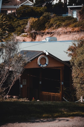 A quaint wooden building nestled among dense greenery features a life preserver with the words 'Canadian Bay Club' displayed prominently at the front. The structure has a small porch with rustic railings, surrounded by trees and shrubs. The background shows a hillside with a mix of vegetation and rooftops of nearby buildings.